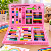 An open pink art kit displays watercolors, crayons, colored pencils, markers, scissors, and a ruler on a wooden table.