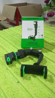 A black and green phone holder disassembled on a green corrugated surface next to its product packaging.
