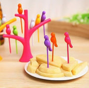 A pink tree holder with colorful bird fruit forks in cheese cubes on a white plate, with a green tree holder in the
