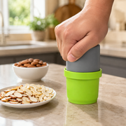 A hand pushes down on a gray plunger inside a green dry fruit slicer, next to bowls of almonds and cashews.