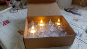 A cardboard box filled with clear acrylic flameless LED tealight candles, some illuminated with warm yellow light.