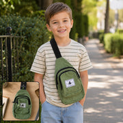 A smiling boy wears an olive green canvas sling bag with a black strap over his shoulder, walking outdoors.