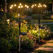 Two solar firefly lights with bright bulbs glow amidst green foliage and colorful flowers in a garden at night.