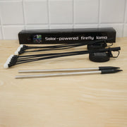 Two solar firefly light units and stakes are displayed in front of a black box on a light wooden surface.
