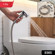 A hand holds a chrome bidet sprayer, spraying water towards a white toilet in a tiled bathroom, with a red 