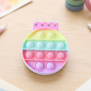 A rainbow pop-it fidget notebook with a pink spiral binding and pastel colored bubbles on a light wooden desk.