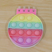 A rainbow pop-it fidget notebook with a pink spiral binding and pastel colored bubbles on a wooden surface.