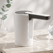 The white and black electric water dispenser pump sits on a marble surface with a glass vase holding eucalyptus branches in