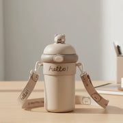 A tan sipper bottle with a bear on top, a 'hello!' inscription, and a patterned strap sits on a desk.