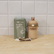 A tan sipper bottle with a bear on top, a patterned strap, and its product box, on a wooden surface.