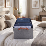 A gray foldable fabric storage basket filled with folded clothes on a bed in a bedroom setting.