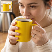 A woman in a cream sweater sips from a bright yellow mug with a white interior, holding it with both hands.