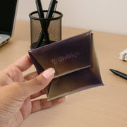 A hand holds the iridescent purple and gold coin pouch, showing its folded design on a wooden desk.