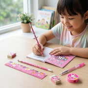 A smiling young girl writing in a small notebook with a pink pencil, surrounded by the matching stationery set on a wooden