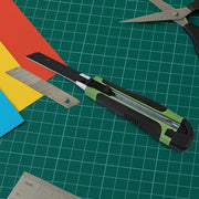 A green and black utility knife with a loose blade and colorful paper on a green cutting mat.