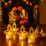 Four golden LED diya tealight candles on a wooden surface with Hindu deities and incense sticks in the background.