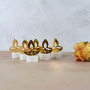 Six golden LED diya tealight candles arranged on a light brown surface with a bouquet of orange flowers on the right.