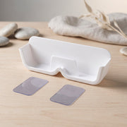 White plastic eyewear holder with two clear adhesive strips on a wooden surface, with decorative stones and fabric in the