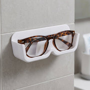 White plastic wall-mounted eyewear holder with tortoise-shell glasses, positioned on a tiled wall next to a mirror.