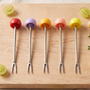 Five stainless steel fruit forks with colorful spherical handles are arranged neatly on a light wooden cutting board.
