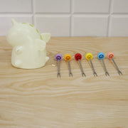 A cream unicorn stand sits beside six colorful fruit forks laid out on a light wooden table against a white tiled wall.