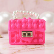 A vibrant pink pop-it purse with a pearl handle and gold clasp sits on a marble surface with blurred white and gold