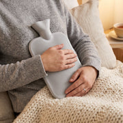 A person in a grey sweater holds a grey ribbed hot water bottle to their chest, with a cream knitted blanket on their lap.
