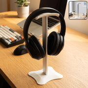 White and silver headphone stand holding black over-ear headphones on a wooden desk with a laptop.
