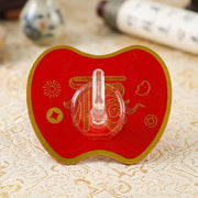 A red apple-shaped hook with gold Chinese characters and a dragon design rests on a patterned tablecloth.