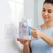 A woman places a clear caddy with shampoo and toothpaste onto a clear wall hook on a white tiled bathroom wall.