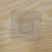 A clear, square self-adhesive hook is centered on a light brown wooden surface with a visible grain.