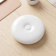 A white, round LED light with a central power button rests on a light wood desk next to a pink mat