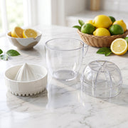 A disassembled white and clear plastic citrus squeezer on a marble counter with lemons and limes in the background.