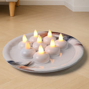 Eight white LED tealight candles with warm yellow flames, arranged on a decorative white plate with feather designs.