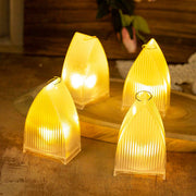 Four transparent arch stripe LED candles glowing brightly on a wooden surface, with blurred green foliage in the background.