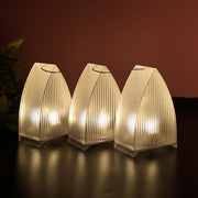 Three transparent arch stripe LED candles glowing on a dark surface, with a subtle green plant in the background.