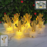 Twelve gold cactus LED tealights with clear textured bases glow warmly on a white surface with green foliage behind.