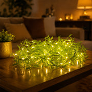 A coiled bundle of bright green leaf string lights with warm white LEDs sits on a wooden table in a dimly lit room.