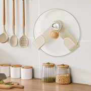 A cream pot lid holder with a wooden knob and chrome center holds a glass lid on a white tiled wall above kitchen counter