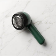 A dark green and black lint remover with a silver mesh head rests on a white marble surface.
