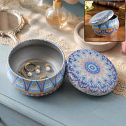 An open blue and orange mandala tin box with a lid next to it, holding gold rings and pearl earrings.