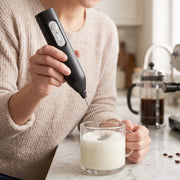 A person in a cream sweater uses a black electric milk frother to create foam in a clear glass of milk on a kitchen counter.