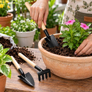 A person uses a small black trowel with a wooden handle to plant a pink flower in a terracotta pot.