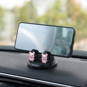 A black phone holder with two pink pig figurines on a car dashboard, holding a smartphone 