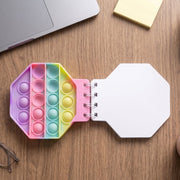 An open pastel hexagon pop it fidget notebook on a wooden desk with a laptop and other office items.