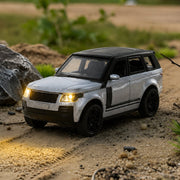 A silver and black die-cast toy SUV with headlights on, casting light on a dirt path with rocks and green foliage.