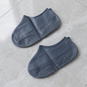 Two dark grey silicone shoe covers with textured soles are neatly placed on a light grey tiled floor.