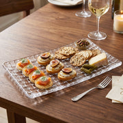 Glass serving tray with a basketweave pattern, laden with appetizers, cheese, and crackers on a wooden table.