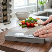 Hands sharpening a silver knife on a gray stone on a wooden cutting board, with fresh vegetables in the background.