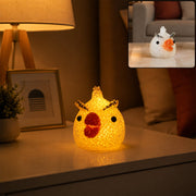 A yellow chick-shaped crystal texture LED lamp glowing on a white nightstand with a small plant and lamp.